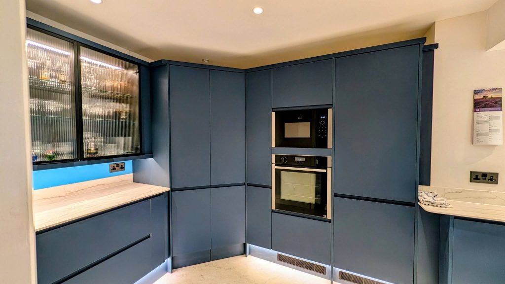Blue handleless kitchen with white worktops and a corner larder and double oven built-it oven housing