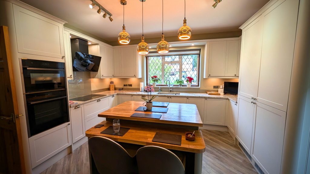 Photograph of a pale Shaker kitchen with a U-shaped layout and central island featuring a two-level timber worktop, with a preparation surface and lower serving area.