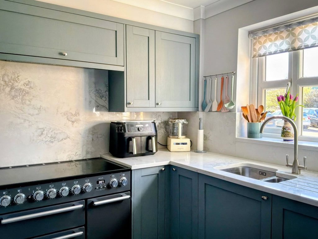 Photograph of a corner of a shaker kitchen in blue-green colour with white quartz worktops, featuring black range cooker and undermounted sink.