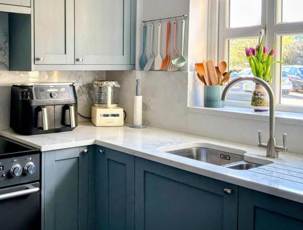Photograph of a corner of a shaker kitchen in blue-green colour with white quartz worktops, featuring undermounted sink.