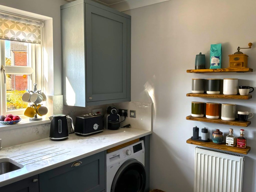 Photograph of a corner of a shaker kitchen in blue-green colour with white quartz worktops, featuring shelves on the wall with jars, and small appliances under the wall cabinet.