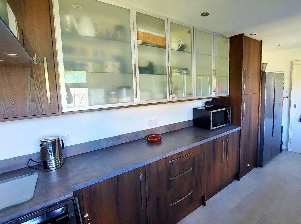 Photo of the side of the galley kitchen in the colour of the warm dark wood grain with sleek brushed steel handles and Caldera laminate worktops. The range cooker at the end has light blue splashback behind and stainless steel finish modern extractor hood above.