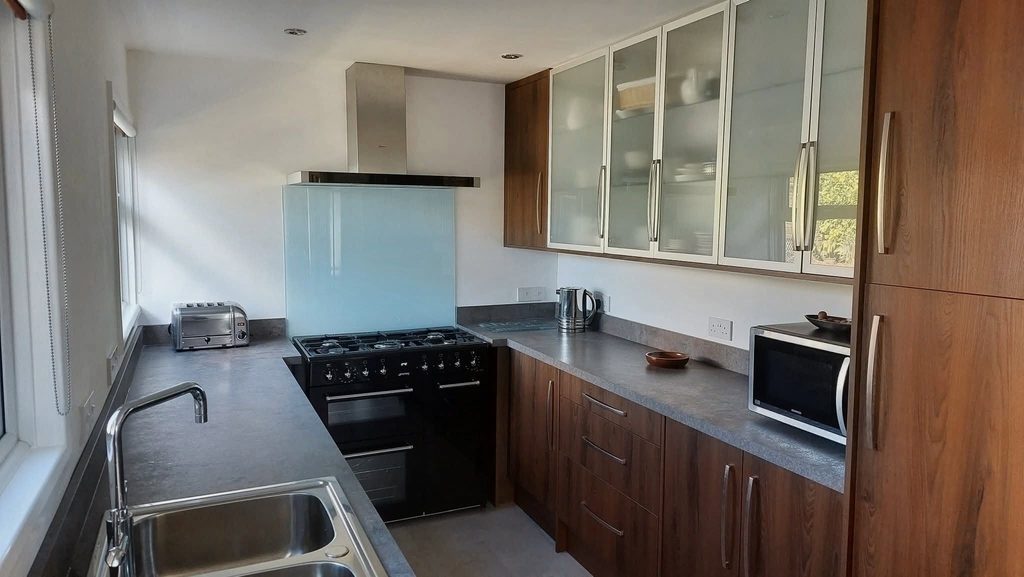 Overall photograph of a galley kitchen featuring dark wood grain cabinets, glazed wall cabinets and a freestanding black gas range cooker. Brushed steel handles and Caldera laminate worktops complete the sophisticated design.
