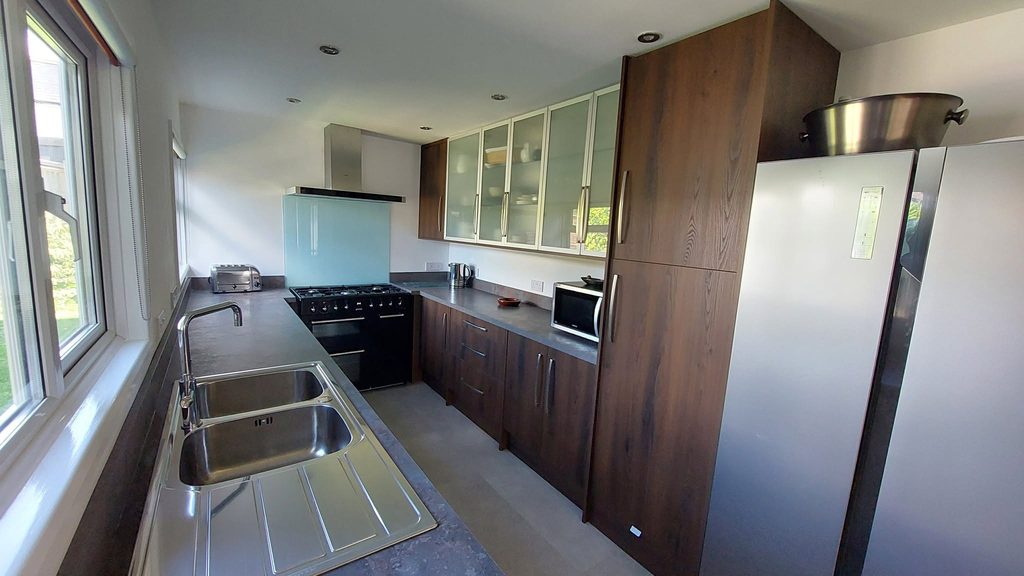 Overall photo of the galley kitchen in the warm colour of the dark grain wood with a freestanding black gas range cooker at the end. Sleek brushed steel handles and Caldera laminate worktops are used to accomplish the deep and sophisticated look.
